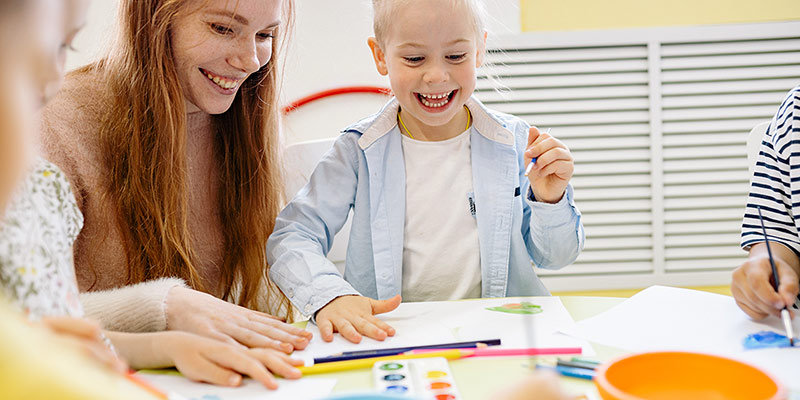 Kleines Mädchen lacht mit einer Kindergärtnerin am Tisch und malt ein Bild