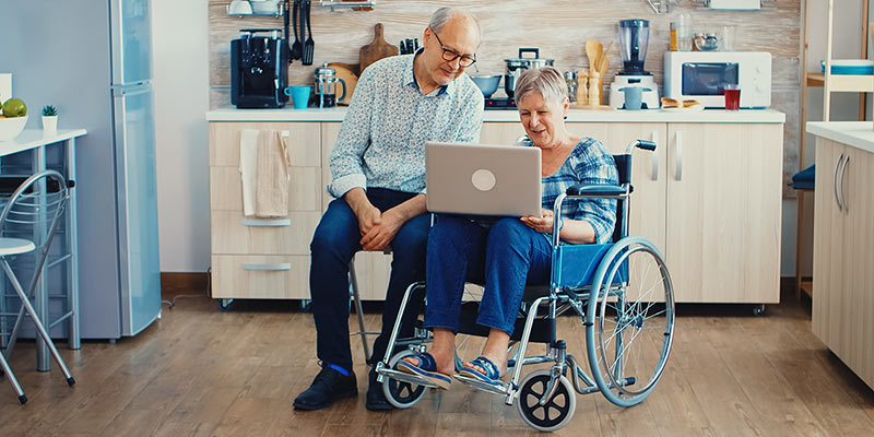 Ältere Frau im Rollstuhl schaut mit ihrem Mann auf einen Laptop