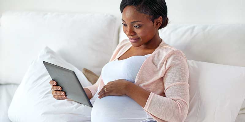 Schwangere Frau sitzt im Bett und hält ein Tablet in der Hand