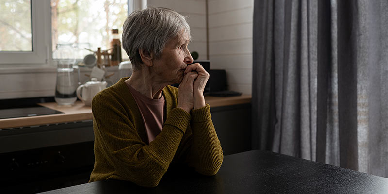 Eine ältere Frau sitzt in der Küche mit zugezogenen Vorhängen und schaut in die Ferne
